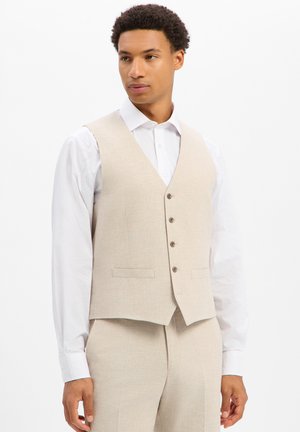 Mann trägt eine beige, knöpfbare Weste über einem weißen Langarmhemd und passenden beigen Hosen vor einem einfarbigen weißen Hintergrund.