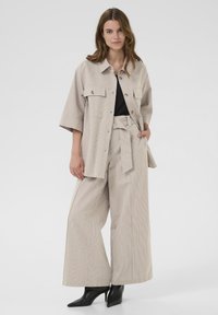 Giacca beige a righe con due tasche frontali e vestibilità rilassata, abbinata a pantaloni a vita alta e gamba larga coordinati e stivali neri a punta.