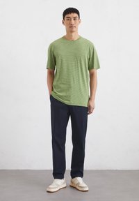 Green striped t-shirt with short sleeves, paired with navy pants. Model wears light-colored sneakers. Background is plain white.