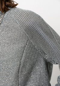 Textuurstoffen in zilver met een ruitpatroon, met een gestructureerd schouderontwerp. Het materiaal heeft een lichte glans en een zachteval.