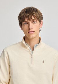Sudadera de cremallera color beige claro, hecha de un tejido suave, que presenta un pequeño logo bordado en el lado izquierdo del pecho y una camisa de cuello debajo.