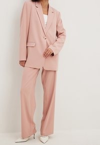 Woman wearing a blush pink oversized blazer and wide-leg trousers paired with white pointed high-heeled shoes standing against a white wall.