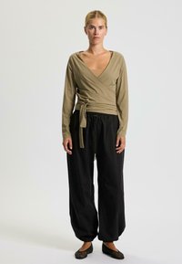 Haut wrap à manches longues en olive clair avec une taille nouée, associé à un pantalon noir ample et des ballerines noires texturées.