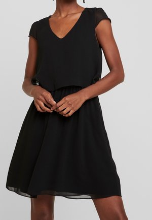 Femme portant une robe noire sans manches en ligne A avec un col en V et une superposition transparente sur un fond clair uni.