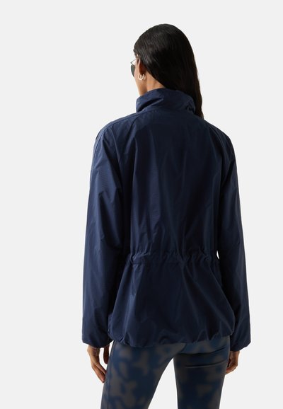 Žena s dlouhými tmavými vlasy, oblečená v tmavě modré bundě a legínách s potiskem, otočená zády k prostému světlému pozadí.