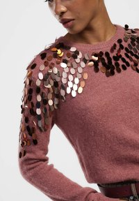 Donna che indossa un maglione a maglia rosa polverosa con grandi paillettes metalliche che abbelliscono le spalle e le maniche superiori.