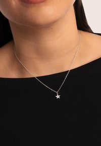 Collana in argento con pendente a forma di stella con superficie texturizzata, indossata su una maglia scura contro uno sfondo neutro.