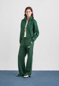 Πράσινο αθλητικό μπουφάν με αντίθετες μπλε ραφές και ψηλό γιακά, συνδυασμένο με ταιριαστά πράσινα αθλητικά παντελόνια με λογότυπο. Τα υφάσματα είναι λεία.