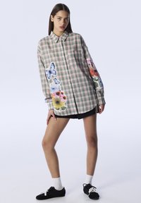 Camicia a maniche lunghe a quadretti verde e rosa, con stampe floreali e di farfalle su un lato, chiusura con bottoni e colletto classico.
