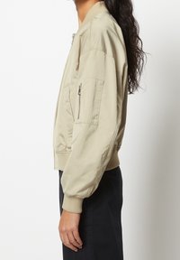 Eine Frau trägt eine beige Bomberjacke mit einem Reißverschlusstaschen an der Ärmelseite und steht seitlich vor einem weißen Hintergrund.
