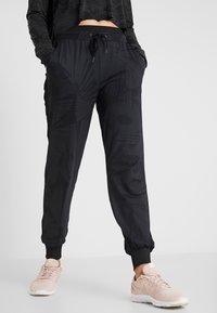Black patterned joggers made of soft fabric with an elastic waistband, side pockets, and tapered cuffs. Worn with light pink athletic shoes.