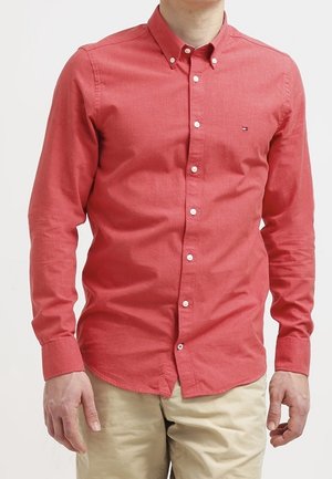 Chemise rouge à boutons fabriquée en tissu texturé avec un col, dotée de boutons blancs et d'un petit logo sur la poitrine gauche. Portée avec un pantalon beige.