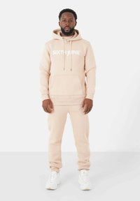 Beige hoodie met "SIXTH JUNE" in het wit gedrukt, met een voorzak en een trekkoord. Gecombineerd met bijpassende beige joggers en witte sneakers.
