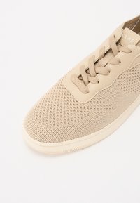 Scarpa sportiva beige con tomaia in rete, punta arrotondata e texture a motivo. Presenta lacci piatti e suola in gomma. Design semplice.