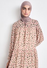 Flytende, blomstrete kjole i lys ferskenfarge med mørke blomsterdesign. Har rund hals, samlet midje og lange ermer. Pyntet med knappdetaljer.