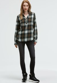 Karierte Hemdjacke in grün, schwarz und weiß mit orangefarbenen Akzenten, kombiniert mit dunklen, slim-fit Jeans und schwarzen Sportschuhen.