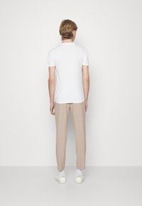 T-shirt bianca a maniche corte abbinata a pantaloni beige sartoriali e scarpe da ginnastica bianche, vista da dietro su uno sfondo semplice.