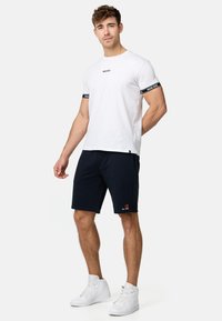 Hvid bomulds t-shirt med sorte detaljer på ærmerne, parret med mørkeblå shorts med et logo. Modellen bærer hvide høj-top sneakers.