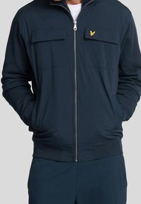 Marineblå zip-up jakke lavet af glat stof, med to brystlommer, ribbede manchetter og et lille gult broderet logo på venstre side.