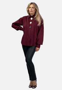 Bordowa koszula z długim rękawem i wysokim kołnierzem; z trzema dużymi srebrnymi guzikami; w połączeniu z czarnymi dopasowanymi spodniami i butami na obcasie.