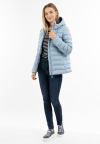 Giacca a vento azzurro chiaro con cappuccio, design trapuntato e chiusura con cerniera, abbinata a una maglietta a righe e jeans scuri.