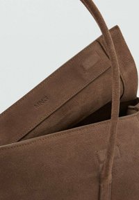 Bolso de ante marrón con textura suave, que cuenta con un asa superior única y una forma rectangular. El interior es parcialmente visible con marca.