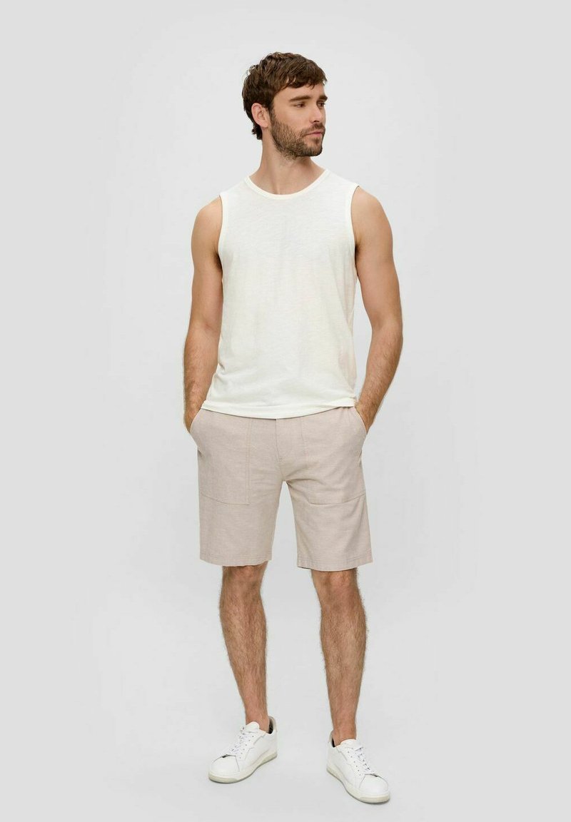 Weißes ärmelloses Tanktop kombiniert mit hellbeigen Shorts. Die Shorts haben zwei Fronttaschen und sind mit weißen Sneakers kombiniert.