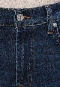 Calças de ganga azul escuro com um botão prateado e costuras laranja. Apresentam fecho de correr, bolsos e um corte clássico de perna reta.