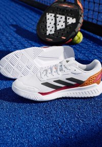 Witte sportschoenen met een mesh bovenwerk, zwarte strepen en oranje accenten; rubberen buitenzool met visgraatpatroon voor grip.