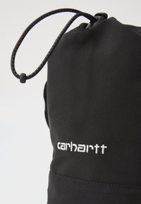 Black drawstring bag made of durable fabric. Features a smooth texture and white embroidered "carhartt" logo. Adjustable closure with a cord.