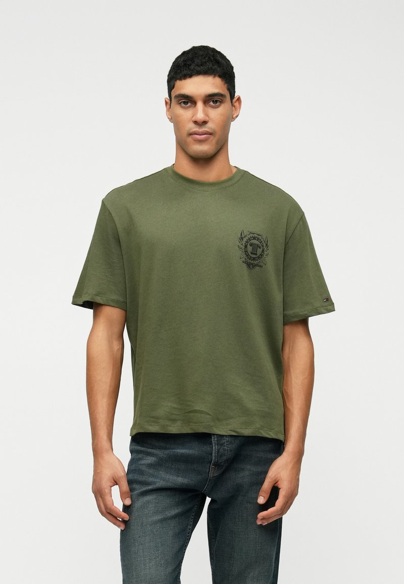 Olivgrön bomullströja med kort ärm, rund hals och ett svart grafiskt emblem på vänster bröst. Avslappnad boxig passform.