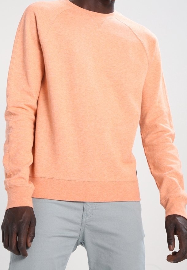 Personne portant un sweat-shirt à manches longues couleur pêche et un pantalon gris clair, debout avec les bras détendus le long du corps sur un fond uni.