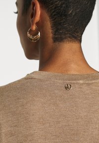 Light brown knitted sweater featuring a round neckline and a small logo detail on the back. Large gold hoop earring visible.