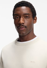 Hellbeiger Pullover mit geripptem Rundhalsausschnitt und dezent auf der Brust gesticktem „BOSS“-Logo. Weiche Textur und minimalistische Design-Details.