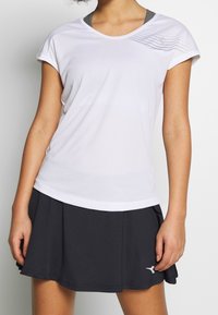 Žena v bílém sportovním tričku s krátkým rukávem a šedým detailem, v černé skládací sukni s malým bílým logem poblíž lemu.