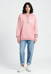 Felpa cappuccio rosa con tasca frontale, con la scritta "HÔTEL DE L'AMOUR" in bianco. Abbinata a jeans azzurri con risvolto e sneaker bianche.