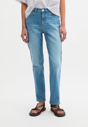 Persoon draagt lichtblauwe rechte jeans met een wit topje ingestopt, staat met ontspannen handen langs het lichaam en donkerbruine sandalen met bandjes.