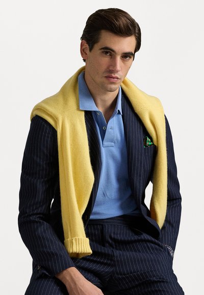 Traje de rayas navy con tela texturizada, camisa de polo azul claro y un suéter amarillo brillante colgado sobre los hombros. Acento de pañuelo verde en el bolsillo.