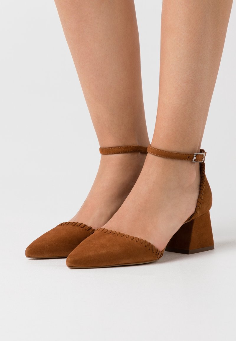 Escarpins en daim marron avec un talon et un bout pointu, dotés d'une bride à la cheville. Ils présentent des coutures le long du bord et un talon carré épais.