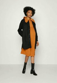 Jaqueta de ganga preta com um colarinho laranja franzido sobre um vestido laranja texturizado. O modelo usa botins pretos brilhantes até ao tornozelo. Look simples e em camadas.