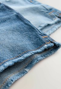 Džinso audinys įvairių mėlynų atspalvių, turintis suirusias kraštines, sustiprintas siūles ir tekstūruotą pynimą, skirtą patvarumui.