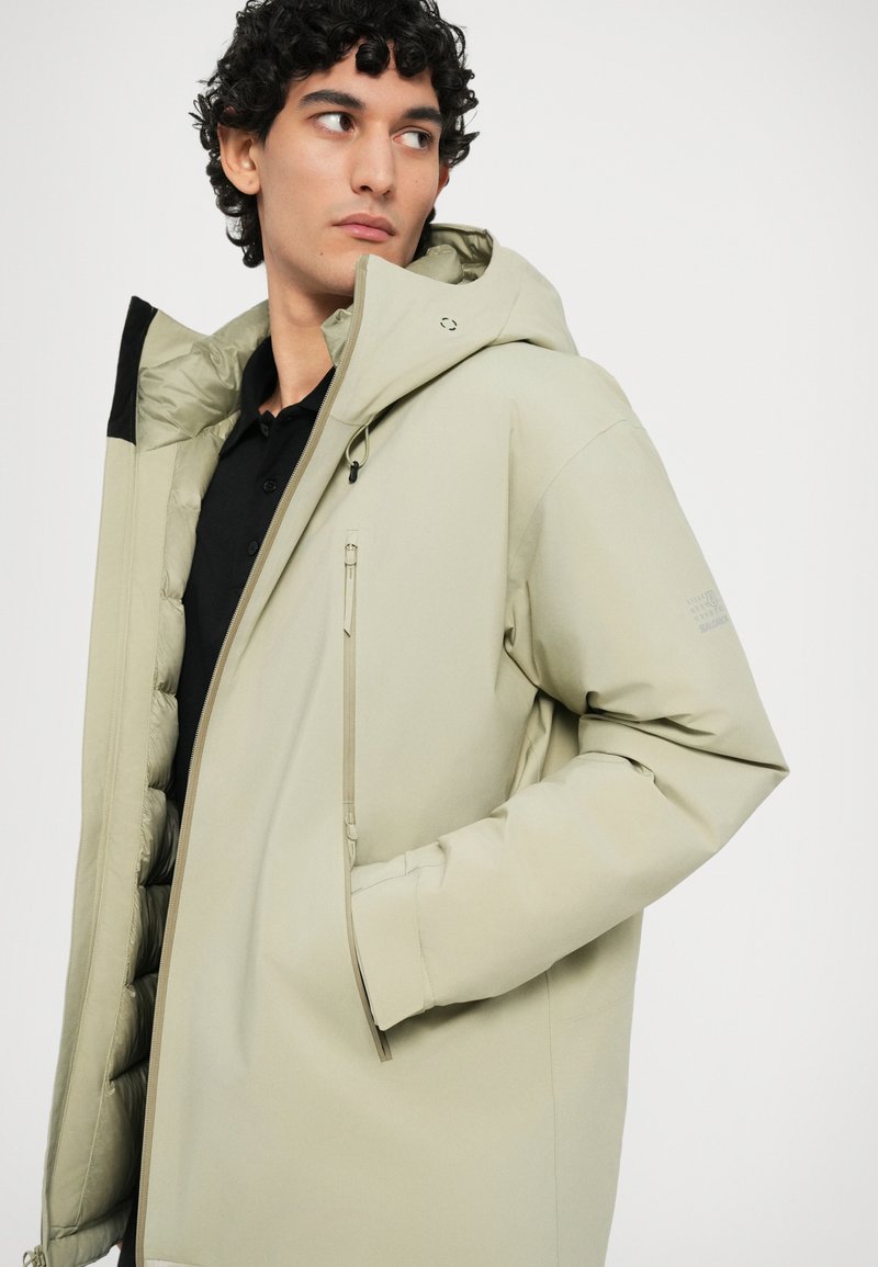 Veste rembourrée beige clair avec une capuche, équipée d'une poche zippée sur la poitrine, en tissu lisse et doublure intérieure texturée. Les accents comprennent des détails noirs.