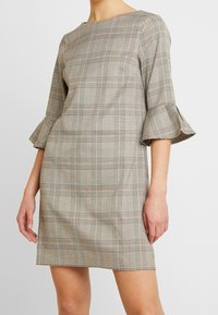 Rutiga klänningen i beige och svart med volangärmar, har rak skärning och rund halsringning, tillverkad av en texturerad tyg.