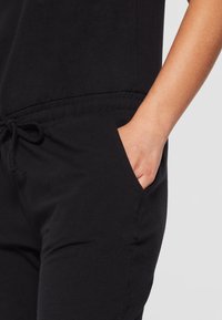 Main humaine reposant dans la poche avant d'un pantalon noir avec une taille élastique et un cordon de serrage.