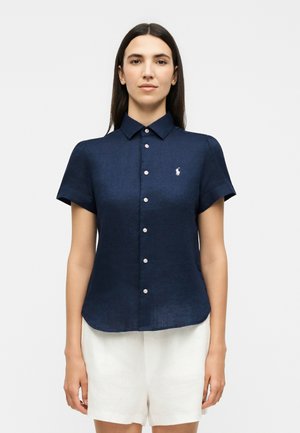 Γυναίκα που φορεί κοντομάνικο μπλε πουκάμισο με κουμπιά και γιακά και άσπρο σορτς, στέκεται σε απλό φόντο.
