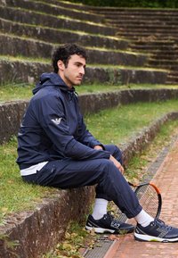 Navy windbreaker with adjustable hood, matching joggers, and black sneakers with white accents, seated on stone steps beside a tennis racket.