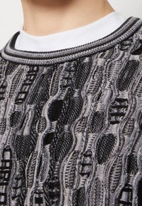 Strickpullover mit einem strukturierten Muster in Schwarz- und Grautönen. Verfügt über einen runden, ribbeligen Ausschnitt in Schwarz und Grau mit einer weißen Unterlage.