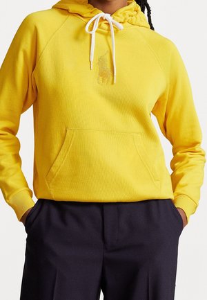 Persoon die een felgele hoodie draagt met witte koordjes en een reliëflogo, gecombineerd met donkerblauwe broek, handen in de zakken.