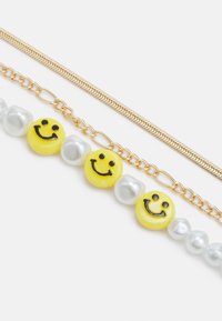 Três pulseiras: a de cima tem uma corrente metálica dourada, a do meio apresenta contas amarelas com sorrisos e a de baixo tem pérolas brilhantes em tons de cinza.
