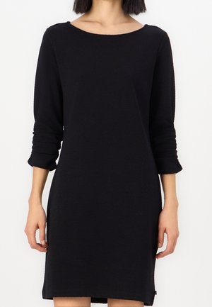 Vrouw draagt een eenvoudige zwarte jurk met lange mouwen en een ronde hals, staand tegen een effen witte achtergrond.